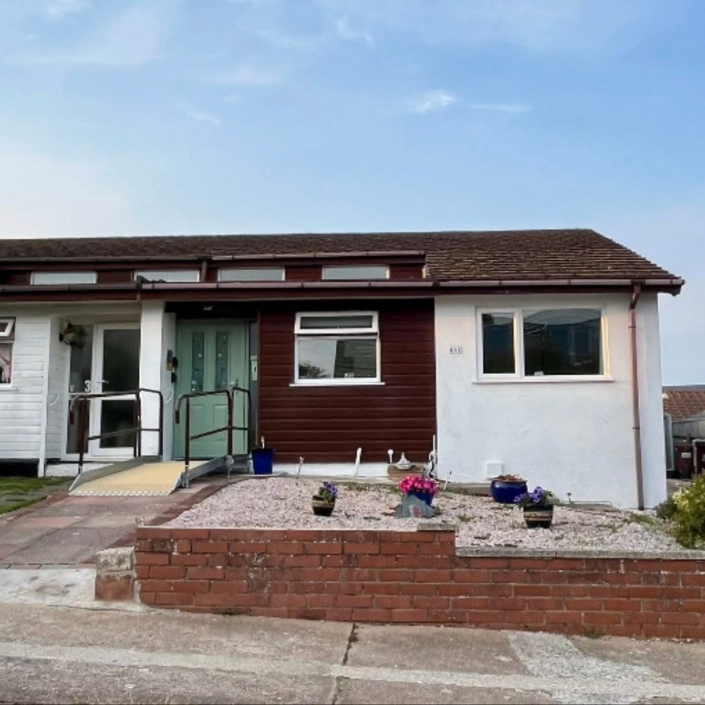 Single-story accessible holiday bungalow with a front wheelchair ramp, handrails, driveway parking, and step-free entry.
