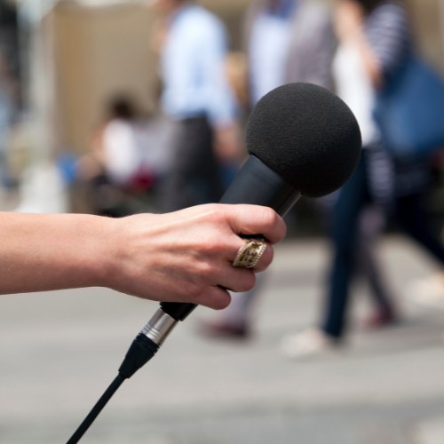 A hand holding a microphone on a city street, preparing to conduct an interview with passersby.