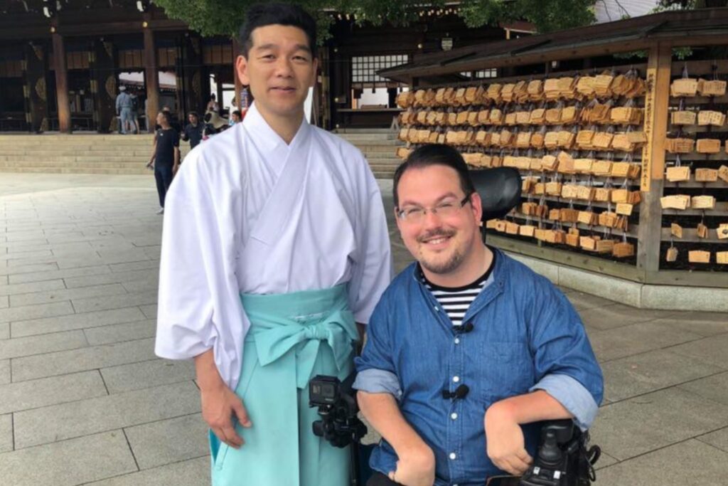 Josh Grisdale, wheelchair user and founder of Accessible Japan, smiling with a Shinto priest at Meiji Shrine, symbolizing Japan’s accessible travel community.