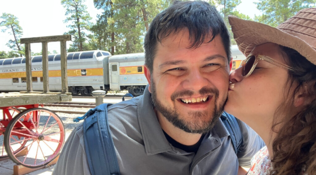 A smiling man with a beard is kissed on the cheek by his wife wearing sunglasses and a sunhat, with the vintage Grand Canyon Railway train in the background at the station.