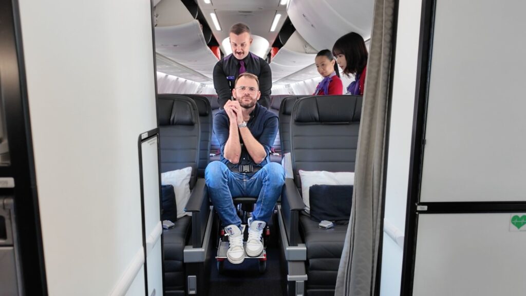 Shane Hryhorec, a wheelchair user, sits in the front row of an airplane cabin while a flight attendant assists him. Two other attendants stand behind, preparing the cabin.