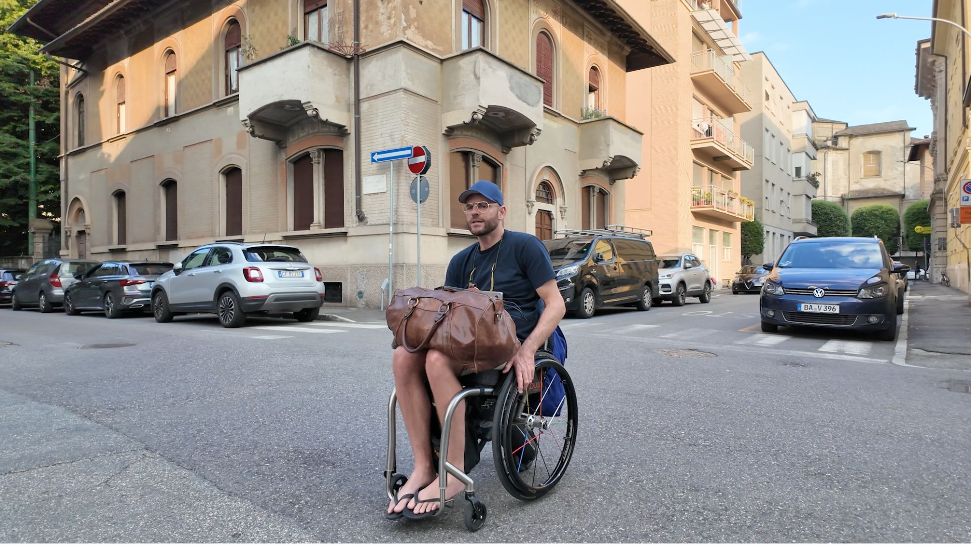 Shane Hryhorec, a wheelchair user, moves along a quiet European street with a large leather travel bag on his lap. Apartment buildings and parked cars line the background.