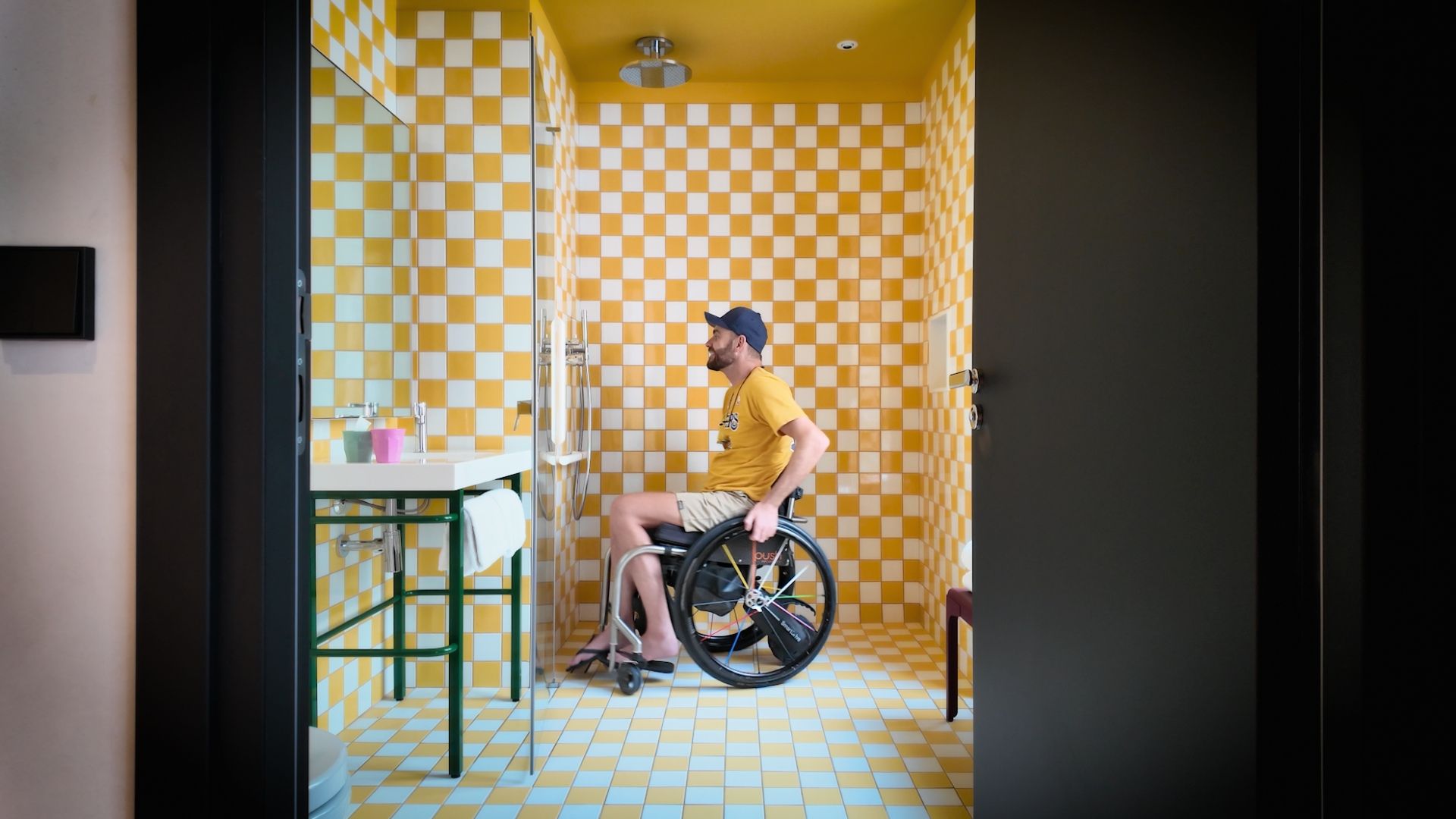 Shane Hryhorec, a wheelchair user, sits in a yellow and white tiled bathroom with a roll-in shower and a sink with space underneath.