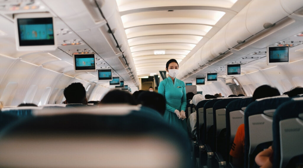 A flight attendant wearing a protective mask stands in the aisle of a passenger aircraft, surrounded by seated travellers, illustrating the challenges wheelchair users face when flying as protections are delayed.