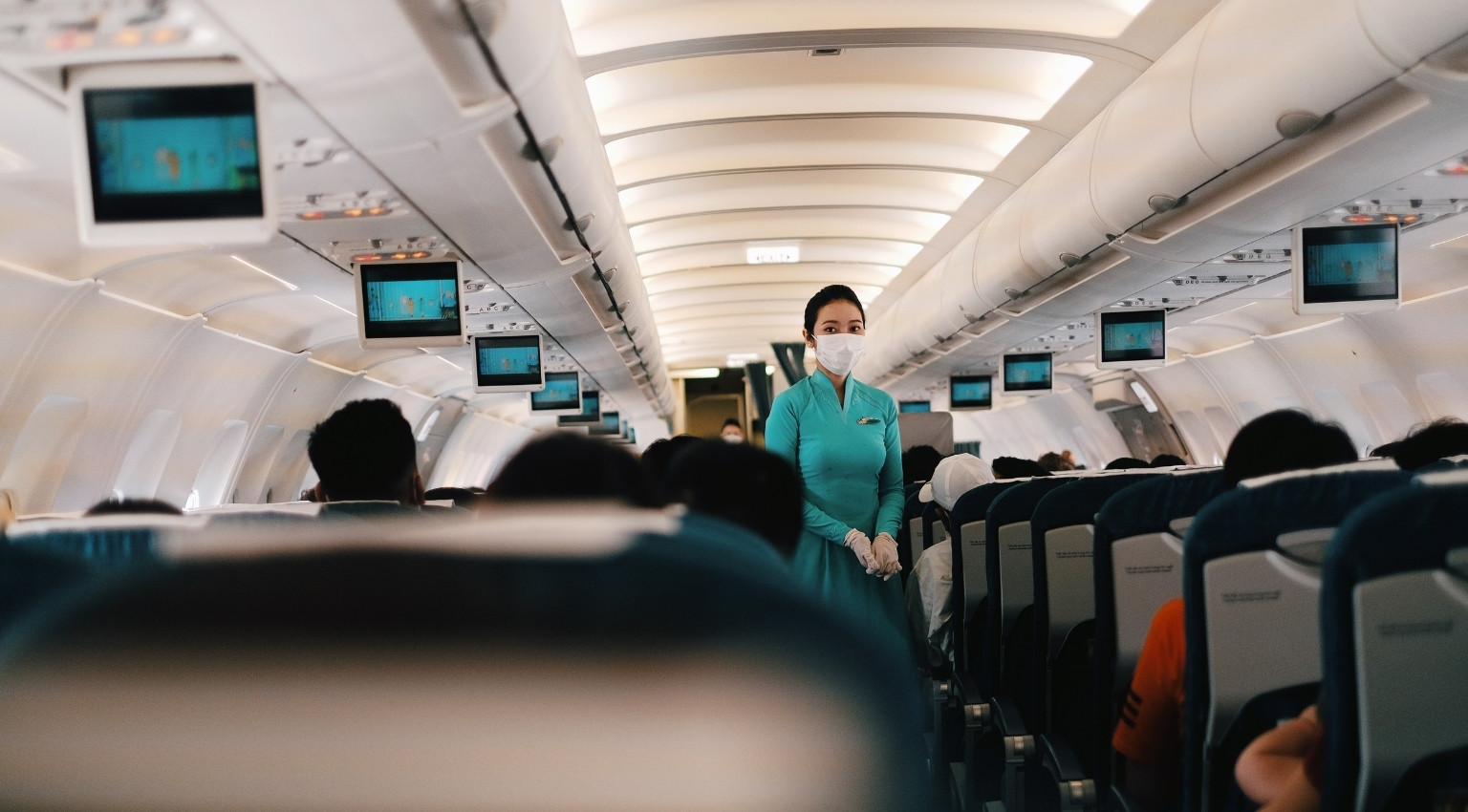 A flight attendant wearing a protective mask stands in the aisle of a passenger aircraft, surrounded by seated travellers, illustrating the challenges wheelchair users face when flying as protections are delayed.