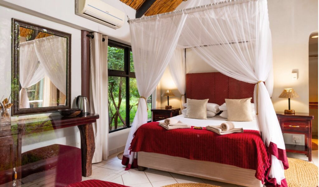 A cozy safari-style bedroom featuring a canopy bed draped with white mosquito netting, red bedding, and beige accent pillows. The room includes wooden furniture, bedside lamps, a mirror, and large windows offering views of the surrounding greenery.