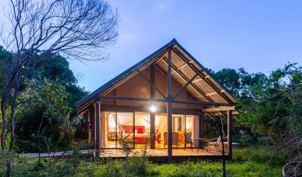 An illuminated forest cabin at Leopard Walk Lodge in KwaZulu-Natal, featuring a thatched roof and large glass doors opening onto a wooden deck. Warm interior lighting reveals a four-poster bed and cozy seating area, surrounded by lush greenery under the evening sky.