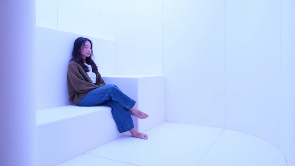 A young lady sits on a large padded step, in a soft padded room called 'The Den' in Changi Airport's Calm Room. The lighting is soft and the environment is tranquil. Perfect for neurodiverse travelers who wish to take a break from the chaos of an international airport.