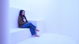 A young lady sits on a large padded step, in a soft padded room called 'The Den' in Changi Airport's Calm Room. The lighting is soft and the environment is tranquil. Perfect for neurodiverse travelers who wish to take a break from the chaos of an international airport.