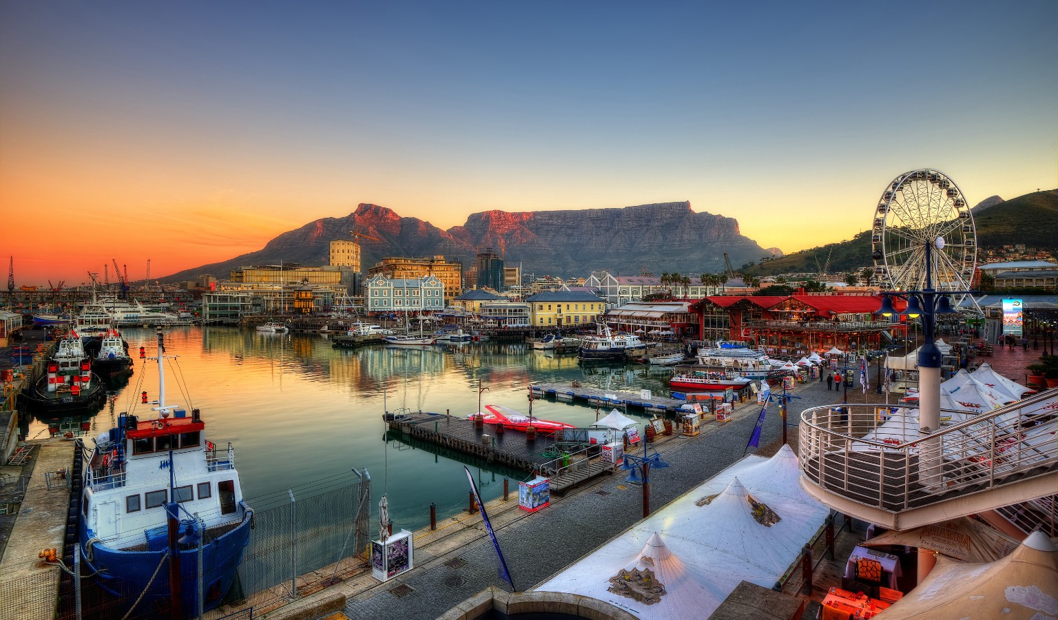 View of Cape Town’s V&A Waterfront at sunset, with Table Mountain glowing in the background. Boats are docked along the calm harbor waters, the Cape Wheel rises on the right, and the waterfront’s colorful buildings and restaurants reflect in the evening light.