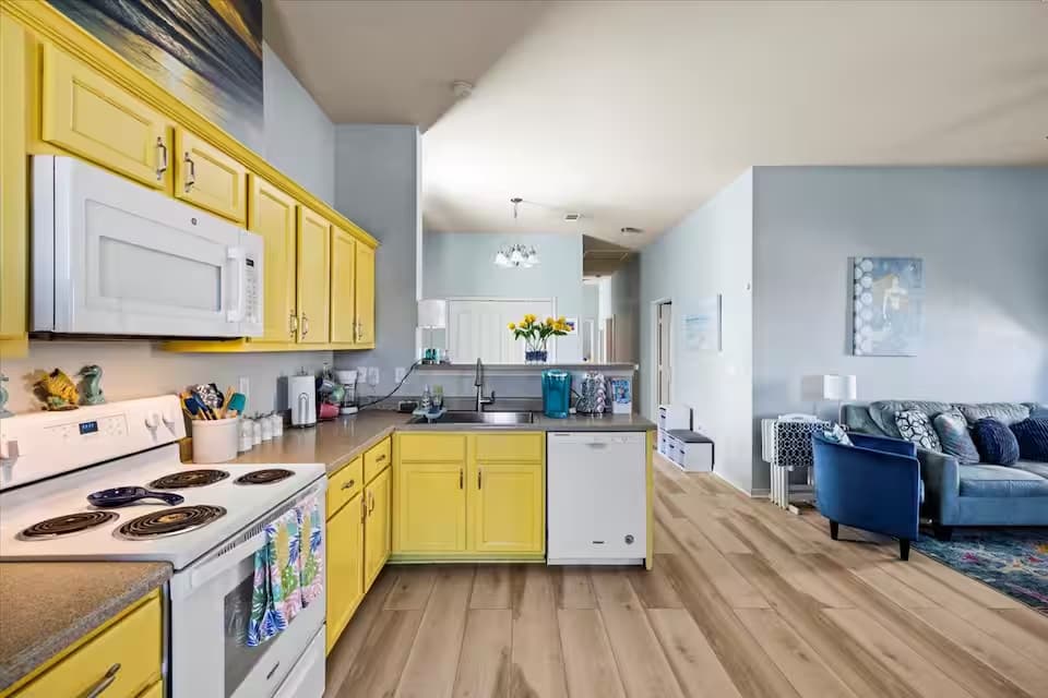 A functional kitchen featuring bold, sunny-yellow cabinetry and light-colored countertops. It includes white appliances: a microwave, a stove/oven, and a dishwasher. A breakfast bar looks out toward the living area.