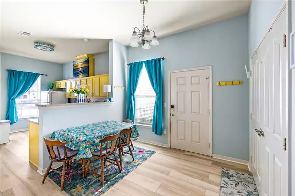 An open-concept dining space with light blue walls and wood-plank flooring. A rectangular table is covered in a colorful starfish-patterned tablecloth and surrounded by four wooden chairs. In the background, a breakfast bar separates the dining area from a kitchen with bright yellow cabinets.