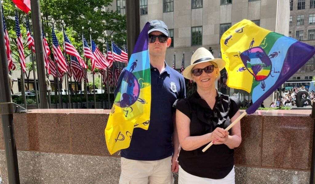 Jeanette Fridh and her son André stand proudly outdoors in New York City, each holding a colorful Disability Flag Precious. The flag features bright blue, green, yellow, purple, pink, and red sections with disability symbols surrounding a globe at its center. Behind them, rows of American flags line the plaza, and sunlight reflects off nearby buildings as people gather in the background.