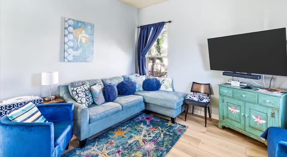 A cozy living area featuring a light blue L-shaped sectional sofa adorned with various blue patterned throw pillows. A vibrant area rug with a large starfish design sits on the wood floor. A teal-colored wooden cabinet sits under a large wall-mounted flat-screen TV.