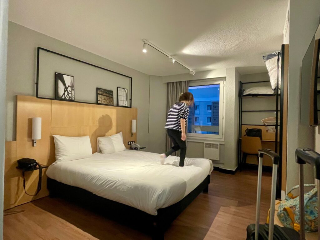 A modern hotel room with a double bed, wooden headboard, framed artwork above it, and a window showing buildings outside at dusk. A person in striped clothing is stepping onto the bed, while suitcases and a small desk area are visible to the side.