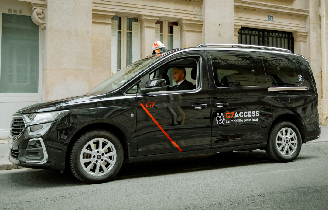 A black taxi-style van parked along a Paris street, marked “G7 Access – La mobilité pour tous,” with a driver visible inside and stone apartment buildings behind it.A black taxi-style van parked along a Paris street, marked “G7 Access – La mobilité pour tous,” with a driver visible inside and stone apartment buildings behind it.