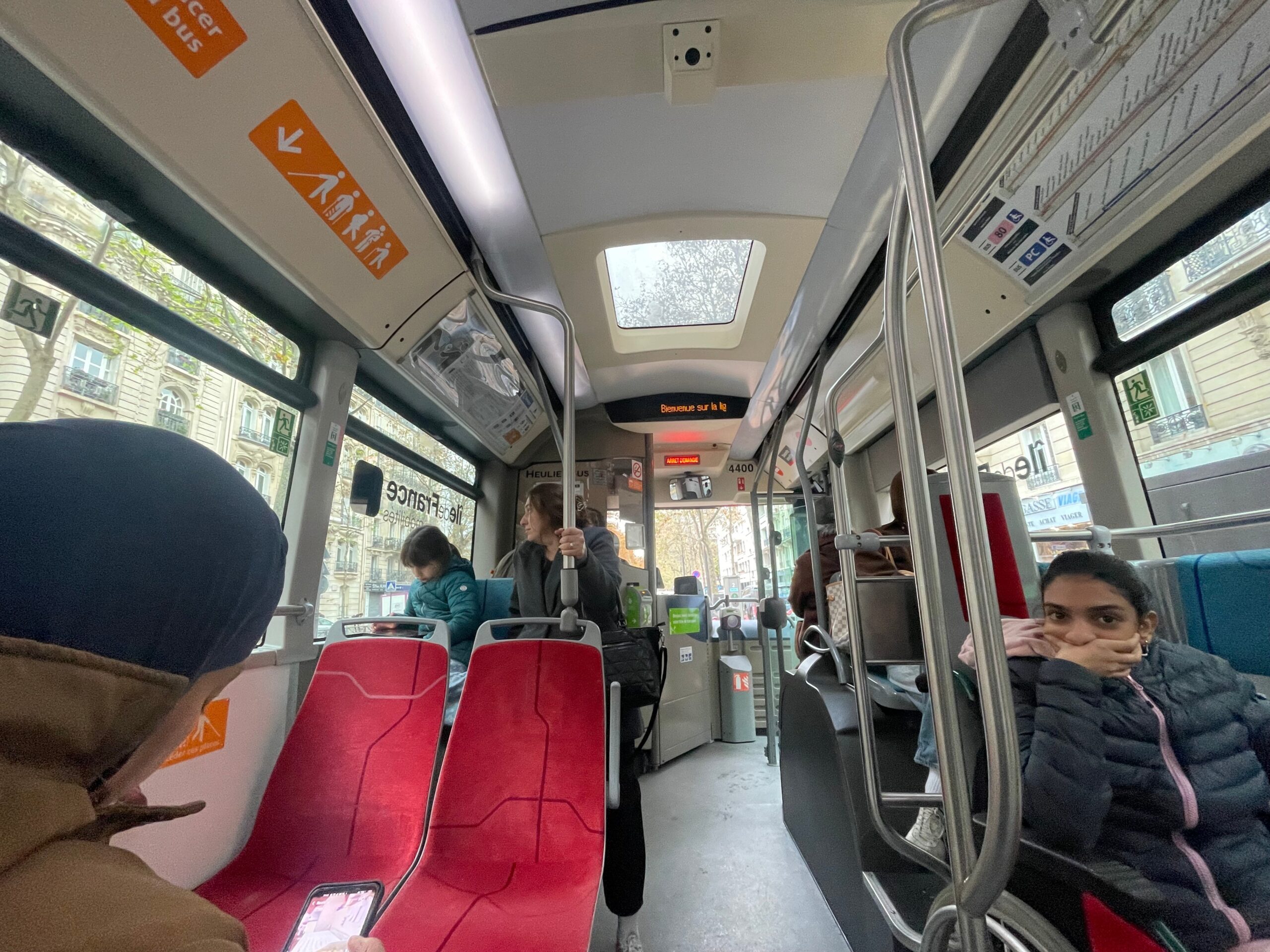 Interior of a Paris city bus with red seats, metal poles, and several passengers seated and standing, including children, while daylight streams through the windows onto the aisle.Interior of a Paris city bus with red seats, metal poles, and several passengers seated and standing, including children, while daylight streams through the windows onto the aisle.