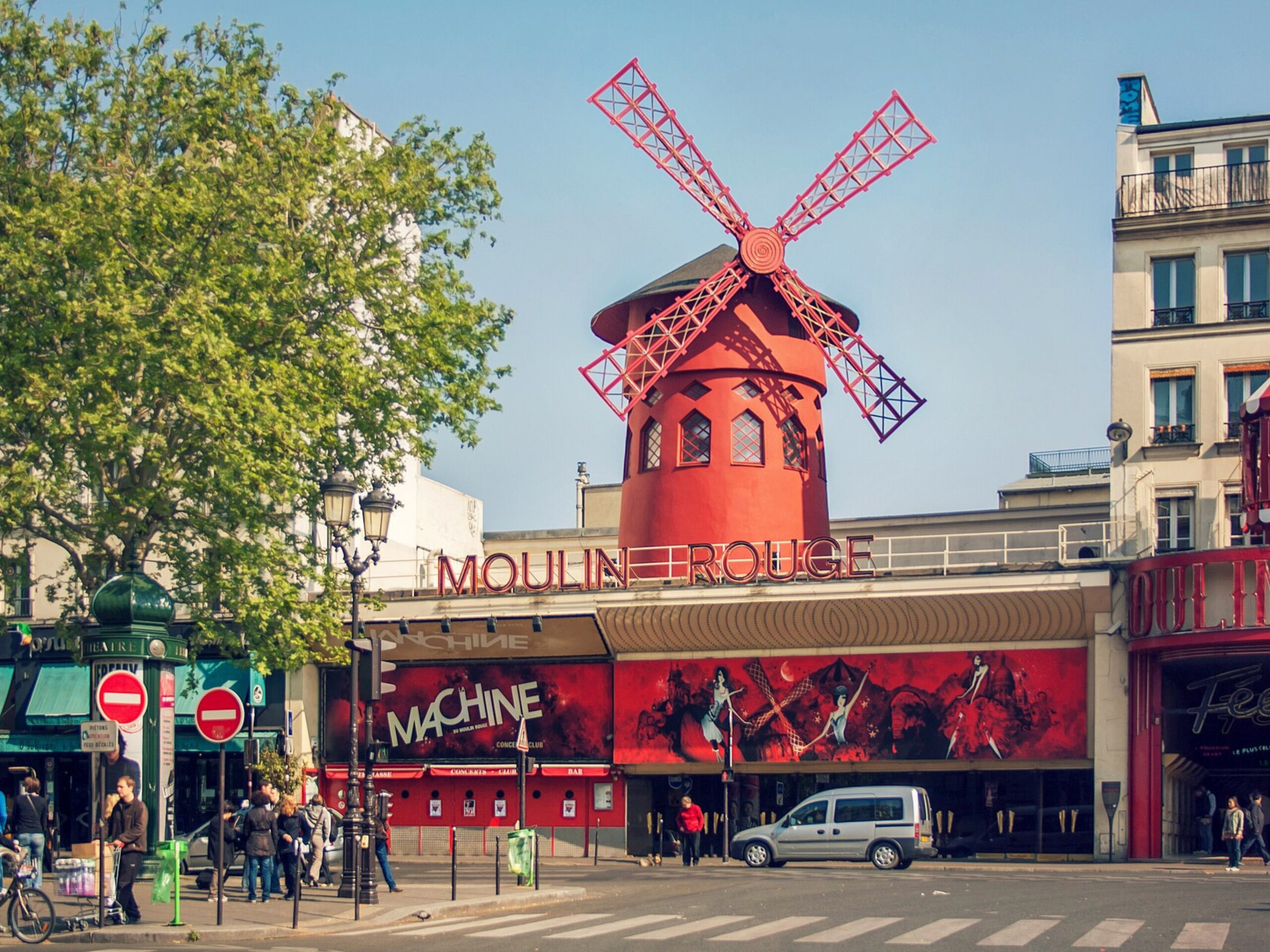 The Moulin Rouge in daylight, with its bright red windmill on the roof, colorful murals across the façade, people gathered on the sidewalk, trees to one side, and city buildings surrounding the famous cabaret.