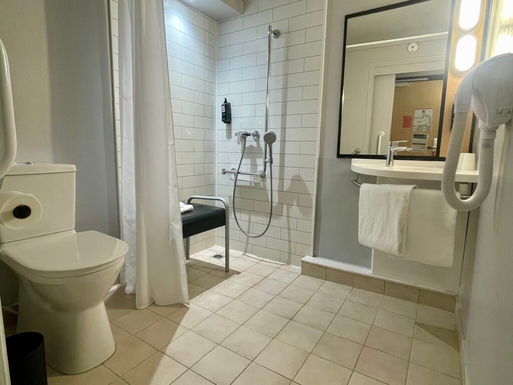 A clean, accessible-style hotel bathroom with tiled floors and walls, a walk-in shower with a handheld showerhead and folding seat, a white shower curtain, a toilet on the left, and a sink with a mirror and hair dryer on the right.
