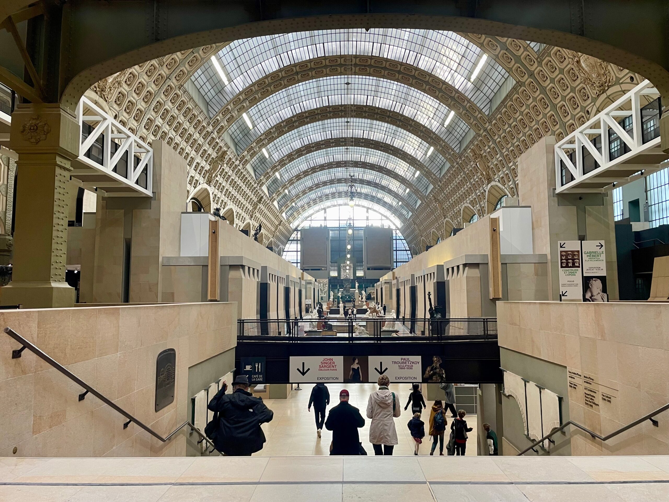 A grand museum hall with a high arched glass ceiling and ornate patterned walls, viewed from the top of a staircase. Visitors walk below toward exhibition areas, with directional signs and sculptures visible in the distance under the long, symmetrical gallery.