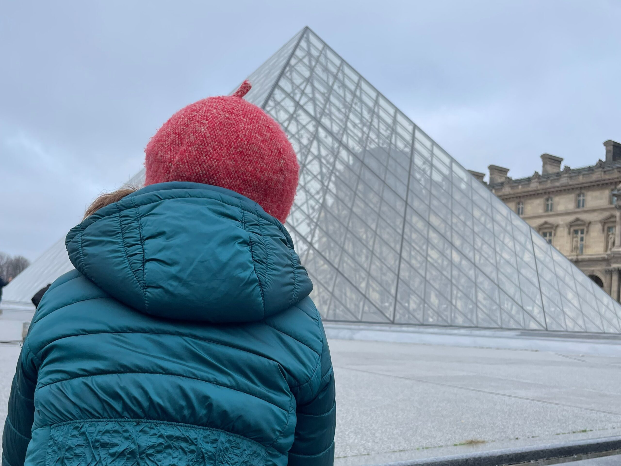 A child wearing a teal puffer jacket and red knit hat stands with their back to the camera, looking up at the glass pyramid of the Louvre Museum under a cloudy daytime sky, with part of the historic palace visible to the side.