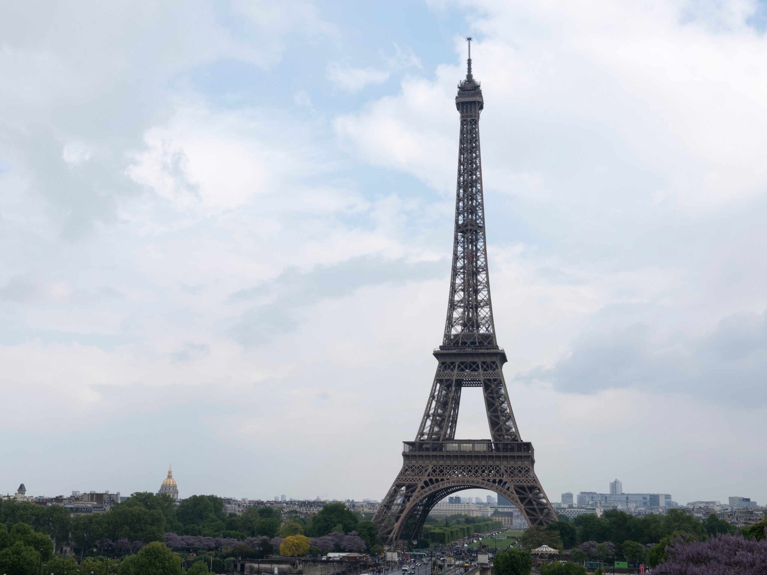 A wide daytime view of the Eiffel Tower rising above Paris, with trees and gardens in the foreground, city buildings stretching across the horizon, and a pale blue sky with scattered clouds overhead.