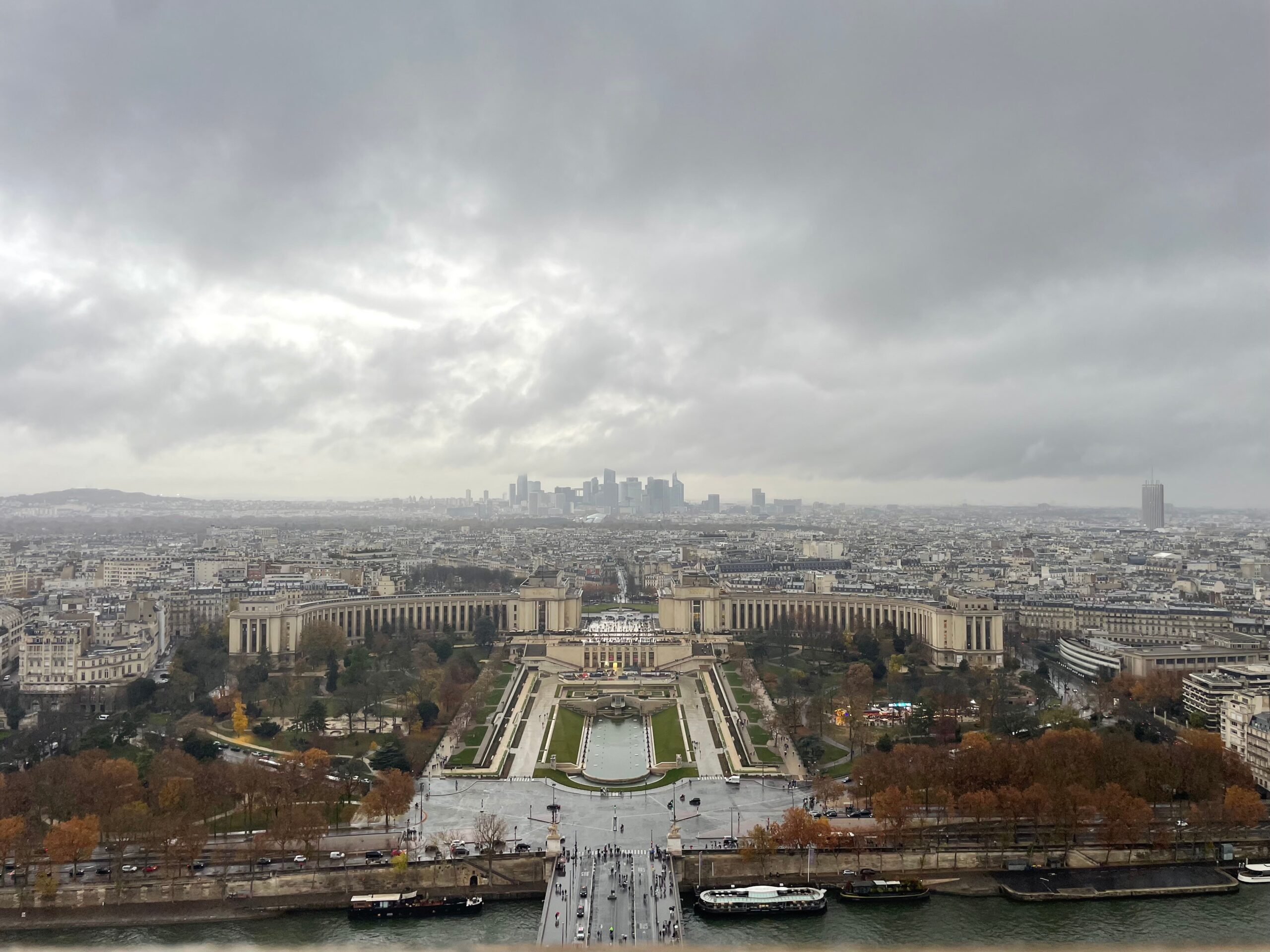 A sweeping elevated view over Paris on a cloudy day, with the Seine River and a bridge in the foreground, the symmetrical gardens and fountains of the Trocadéro below, and the distant skyline of La Défense rising on the horizon.