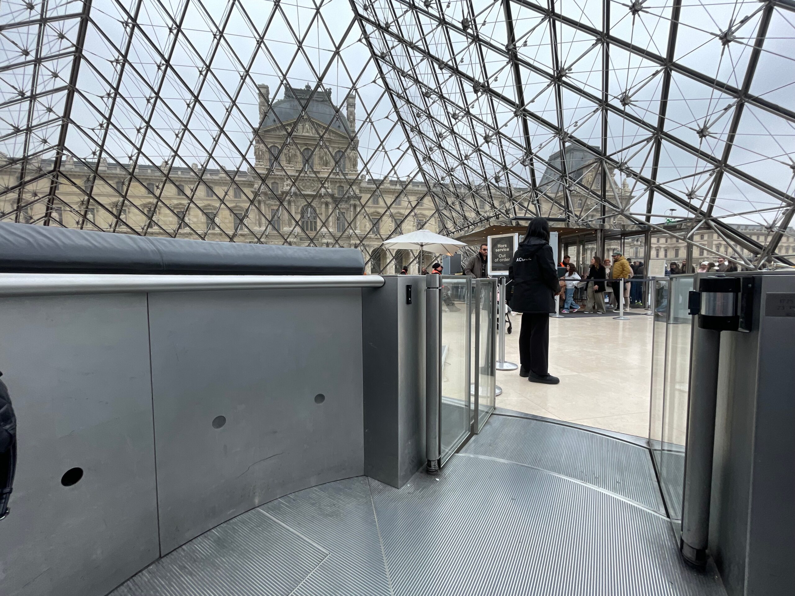 Inside the Louvre’s glass pyramid, metal framing forms a crisscross pattern overhead while visitors stand in line near a security checkpoint. Glass barriers and turnstiles are in the foreground, with the historic museum buildings visible through the pyramid walls.