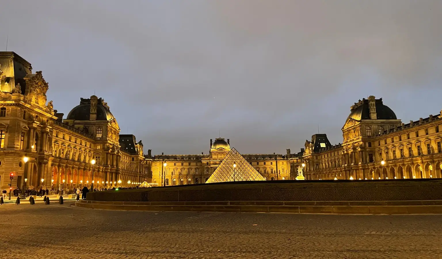 Feature image for 'Paris in a Wheelchair: Our Three Day Itinerary in the City of Love'. A wide nighttime view of the Louvre Museum courtyard in Paris, with the illuminated glass pyramid centered in front of the grand historic palace buildings, warm golden lights glowing along the façades, and a cloudy evening sky overhead.