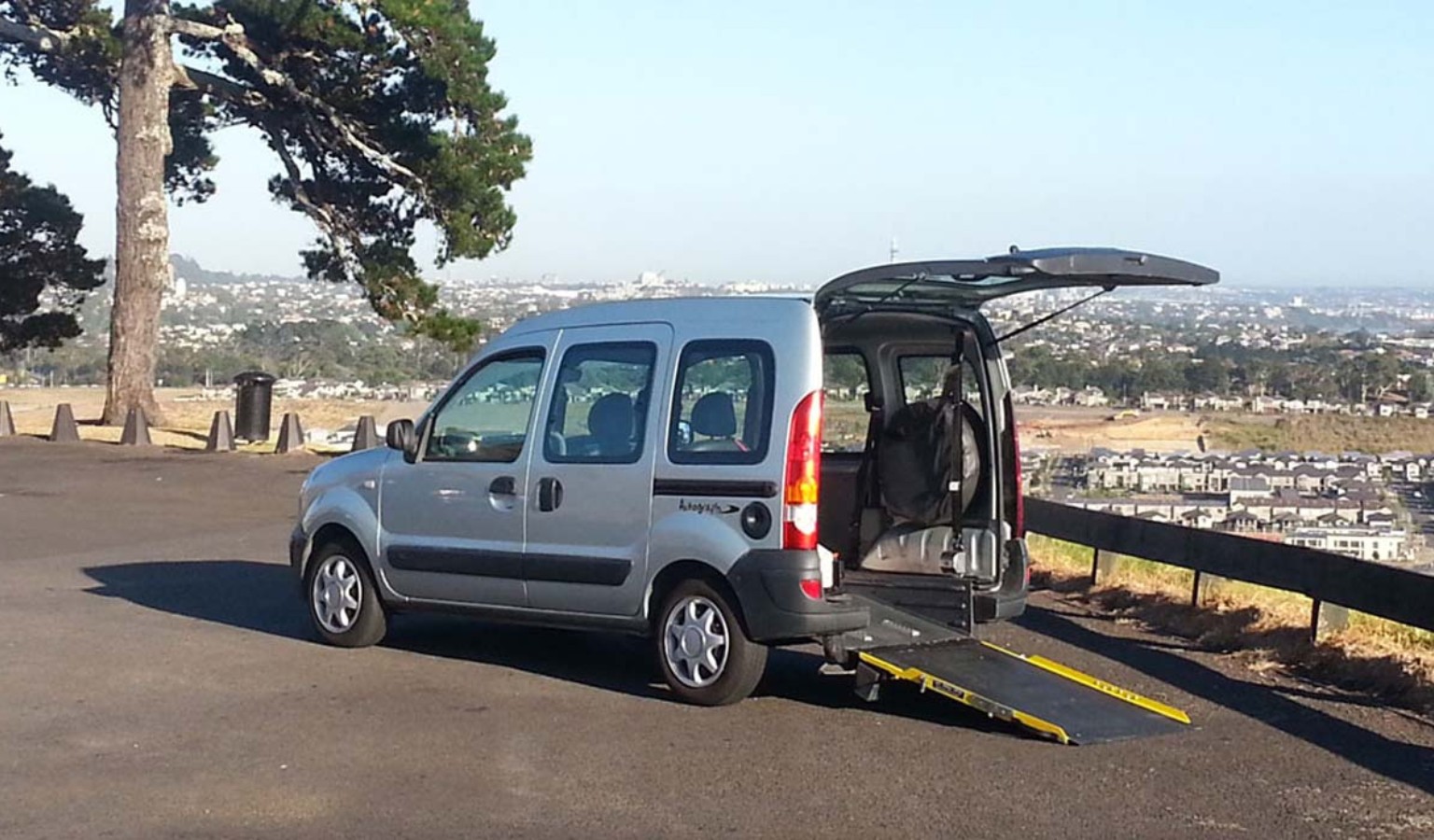 A silver wheelchair-accessible van is parked on a paved overlook with its rear hatch open and a black-and-yellow ramp extended to the ground. Inside the back of the van, a wheelchair is visible. A large tree stands to the left, and beyond the railing in the background is a sprawling cityscape under a clear sky.