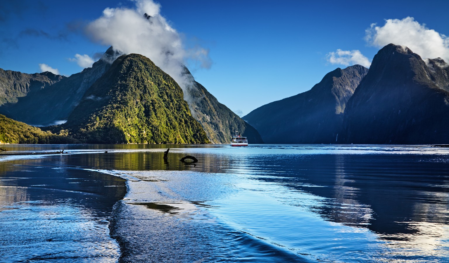A dramatic fjord scene shows steep, forested mountains rising on both sides of calm blue water under a clear sky, with clouds clinging to the peaks. A small red-and-white boat cruises in the distance, while ripples in the foreground reflect the mountains and sunlight.