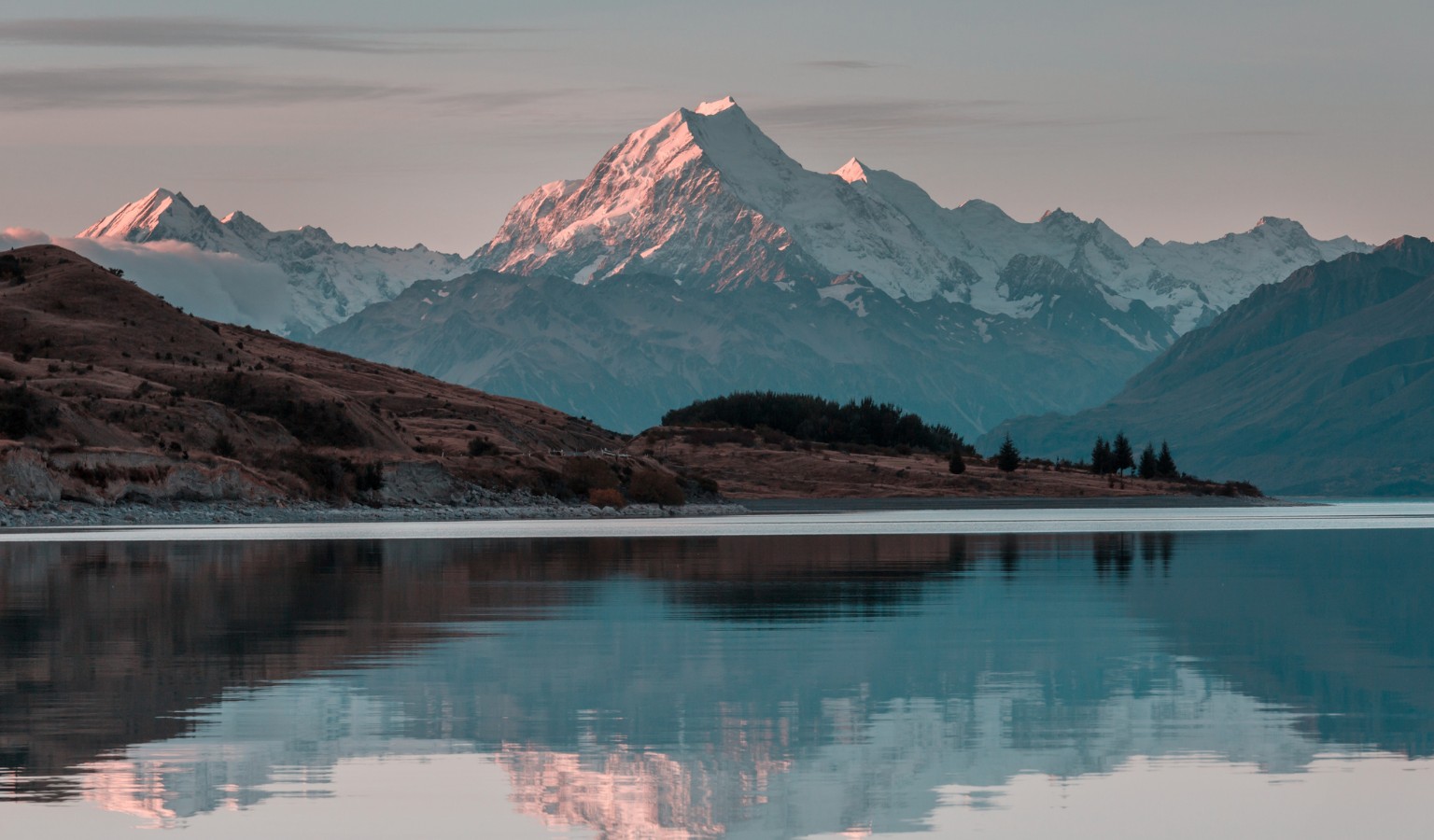 A tranquil lake reflects snow-capped mountains at dusk, with the highest peak glowing pink from the setting sun. Low hills and sparse trees line the shoreline in the foreground, while layered mountain ridges fade into the distance beneath a pale evening sky.