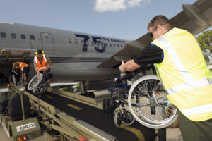 Airport ground crew wearing orange and yellow high-visibility safety vests unload multiple folded wheelchairs from a baggage conveyor belt alongside a silver commercial aircraft. The plane displays "75th Anniversary" lettering on its fuselage. The scene takes place on a sunny tarmac with blue skies and trees visible in the background.