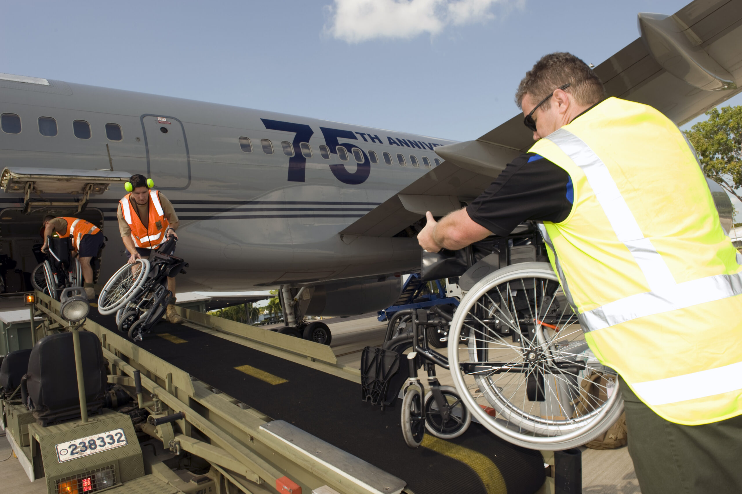 Airport ground crew wearing orange and yellow high-visibility safety vests unload multiple folded wheelchairs from a baggage conveyor belt alongside a silver commercial aircraft. The plane displays "75th Anniversary" lettering on its fuselage. The scene takes place on a sunny tarmac with blue skies and trees visible in the background.