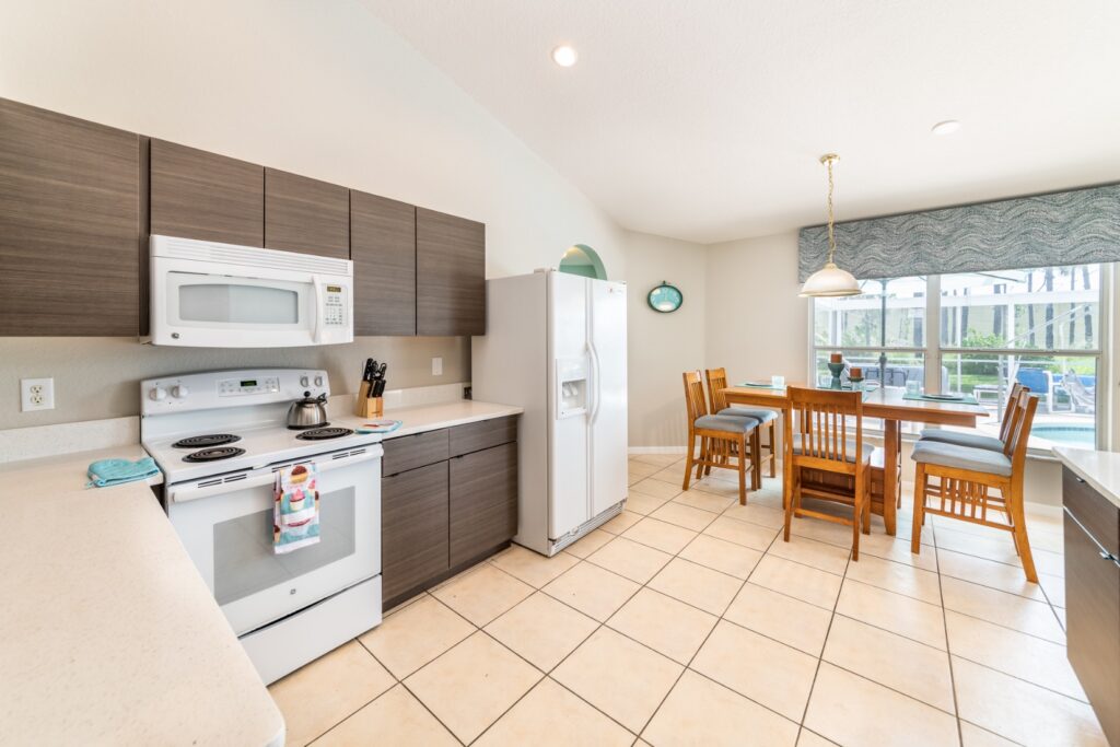 Open-plan kitchen and dining area with dark wood cabinetry, white appliances including electric hob, microwave and large fridge-freezer, generous worktop space, and a six-seat dining table with a pool view through the window.