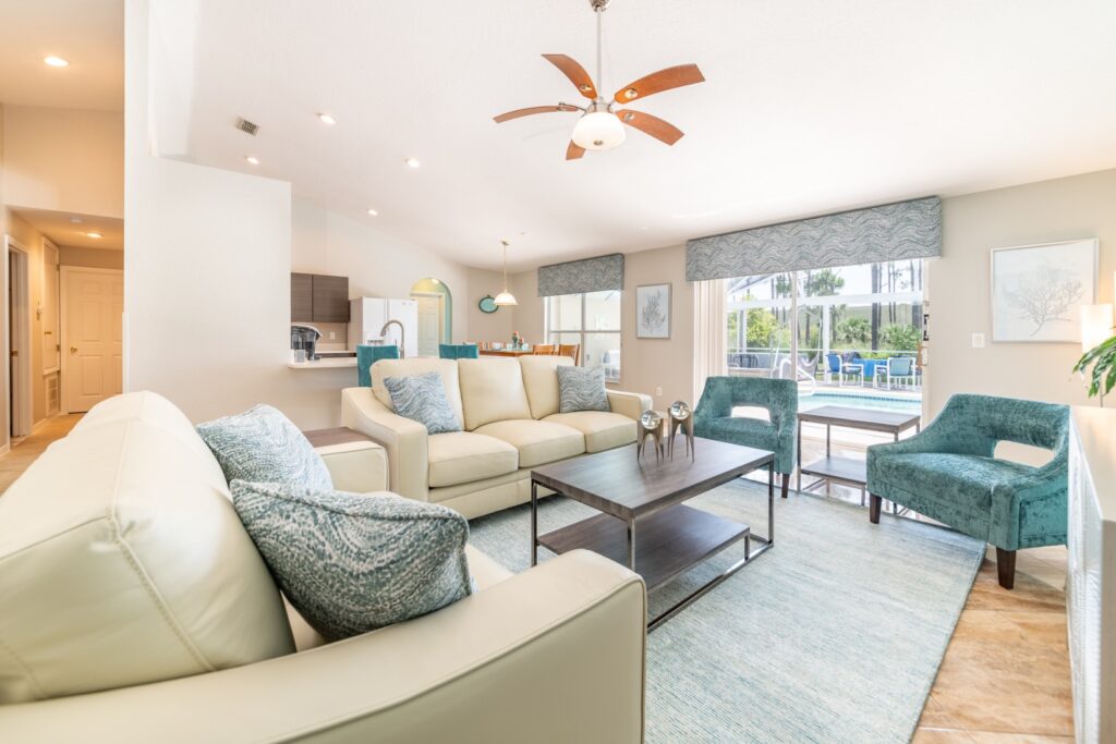 Spacious open-plan living room with cream leather sofas, teal accent chairs, a ceiling fan, and large sliding doors leading directly to the pool area — with the kitchen and dining room visible in the background.