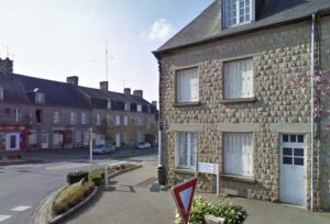 Quiet street corner in Sourdeval, Normandy, showing the traditional stone-built property on a peaceful town square with nearby shops visible in the background.