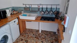 Wheelchair-accessible kitchen with open-under sink and hob at lowered height, wooden worktops, hanging pan rack, dishwasher, washing machine and colourful mosaic tile floor.