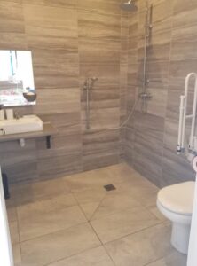 Spacious wetroom with large-format wood-effect tiles, open roll-in shower with rainfall head and handheld attachment, drop-down grab rail, WC, and wheelchair-accessible sink.