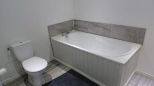 Clean family bathroom with a full-size white bath with grey tiled surround, white panelled bath front, and toilet on wood-effect flooring.