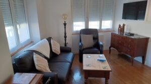 Bright lounge with large windows, a dark leather sofa and armchair with cushions, a wooden coffee table, antique dresser and wall-mounted TV.