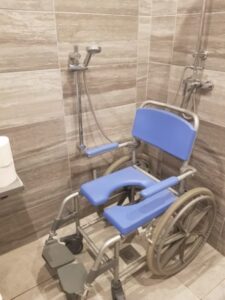 Blue commode shower wheelchair with footrests positioned in the wetroom beside a wall-mounted grab rail and handheld shower head.