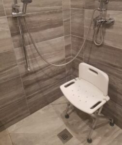 Accessible shower area with wall-mounted grab rail, handheld shower head, and a white fold-down shower chair on a non-slip tiled floor.