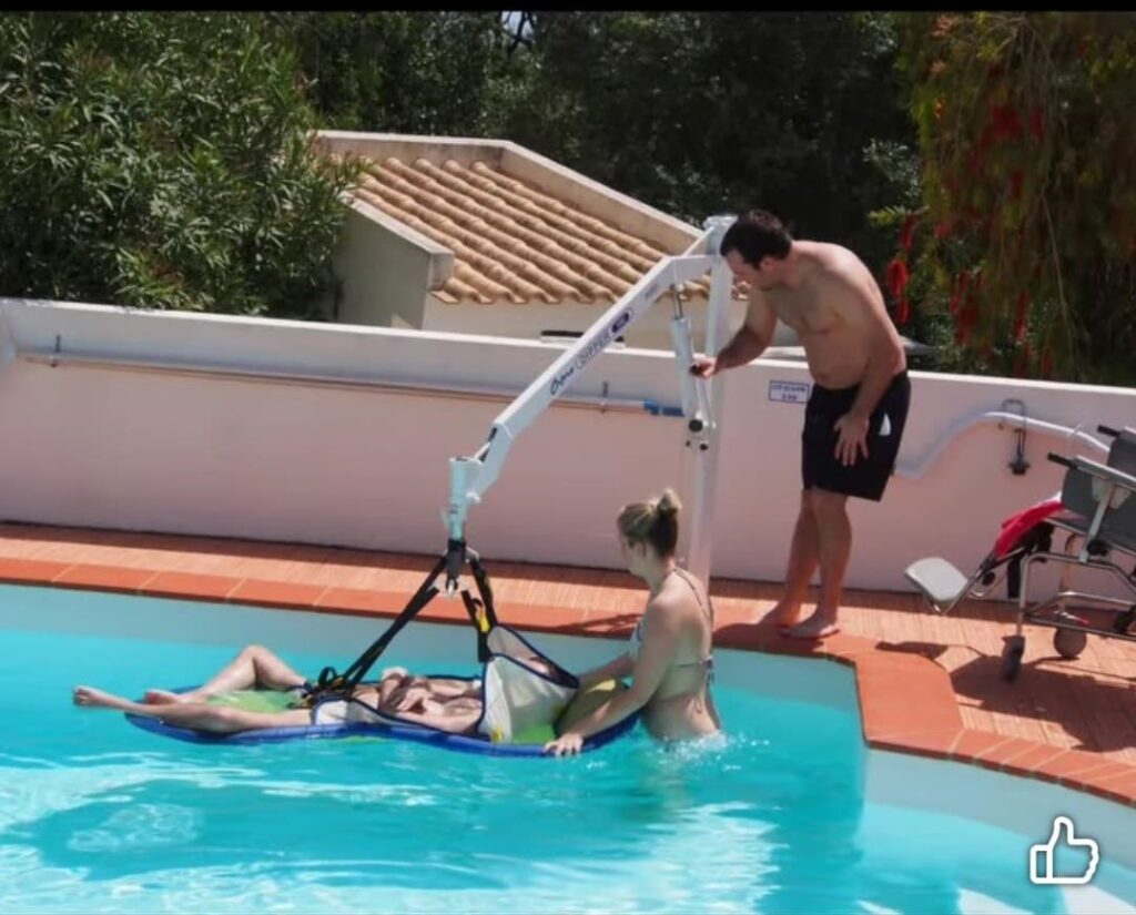 A man stands on a poolside deck operating a white mechanical pool hoist. The hoist is lowering a person supported by a blue and white sling into a clear blue swimming pool, while a woman in the water assists with the positioning.