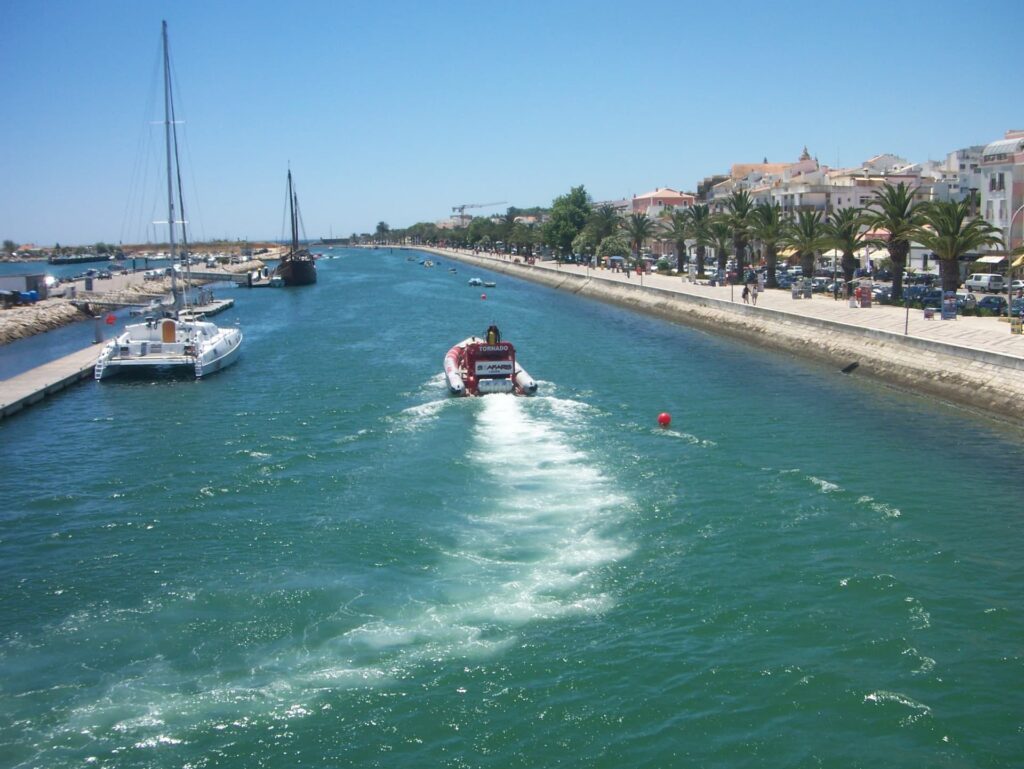 A high-angle view of a wide sea canal in Lagos. A red and white "Tornado Safaris" speedboat creates a long white wake as it travels down the center of the turquoise water. The right bank features a paved promenade lined with palm trees and white buildings under a clear blue sky.