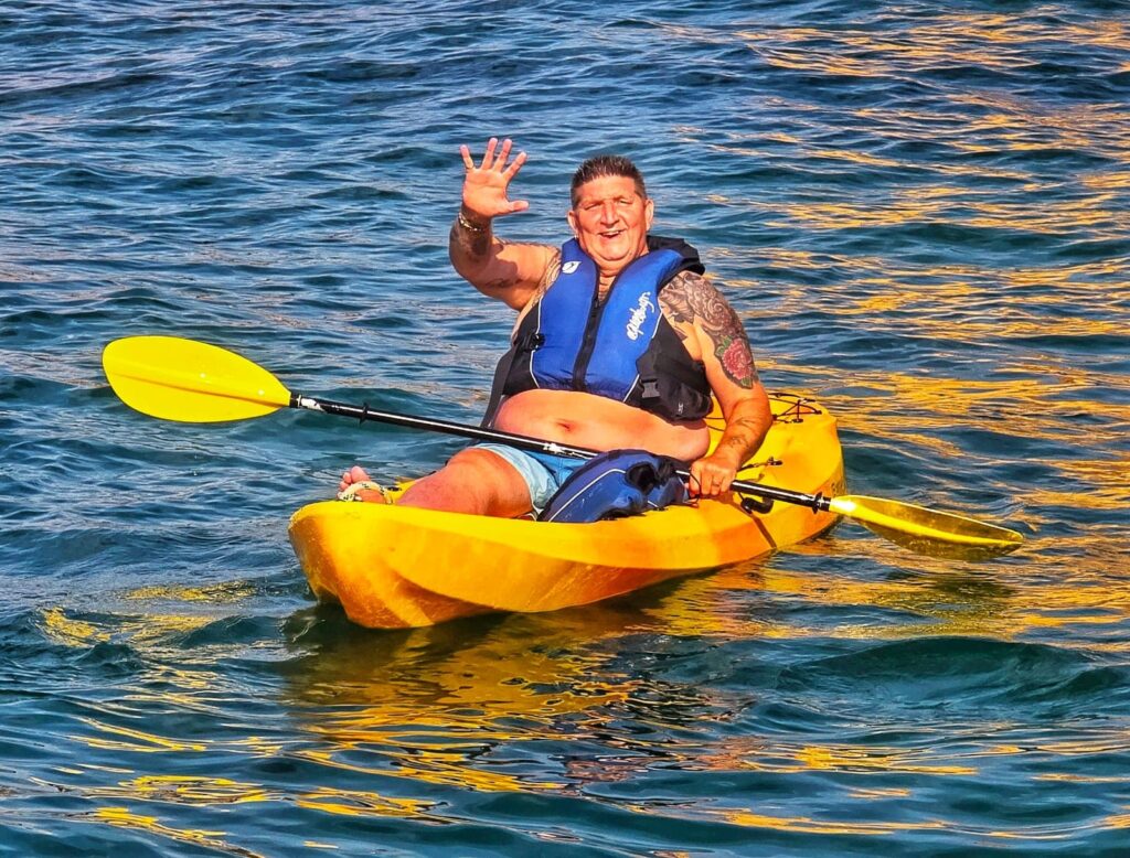 A man wearing a blue life vest sits in a bright yellow kayak on rippling blue water. He is smiling and waving with one hand while holding a yellow-bladed paddle.