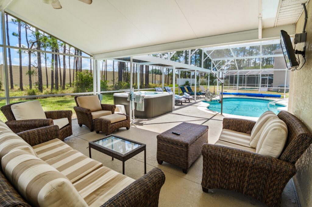 Spacious screened lanai with large wicker sofa, armchairs and coffee tables overlooking a private outdoor pool and hot tub, with ceiling fan and wall-mounted TV. Sunloungers and a pool hoist are visible poolside.