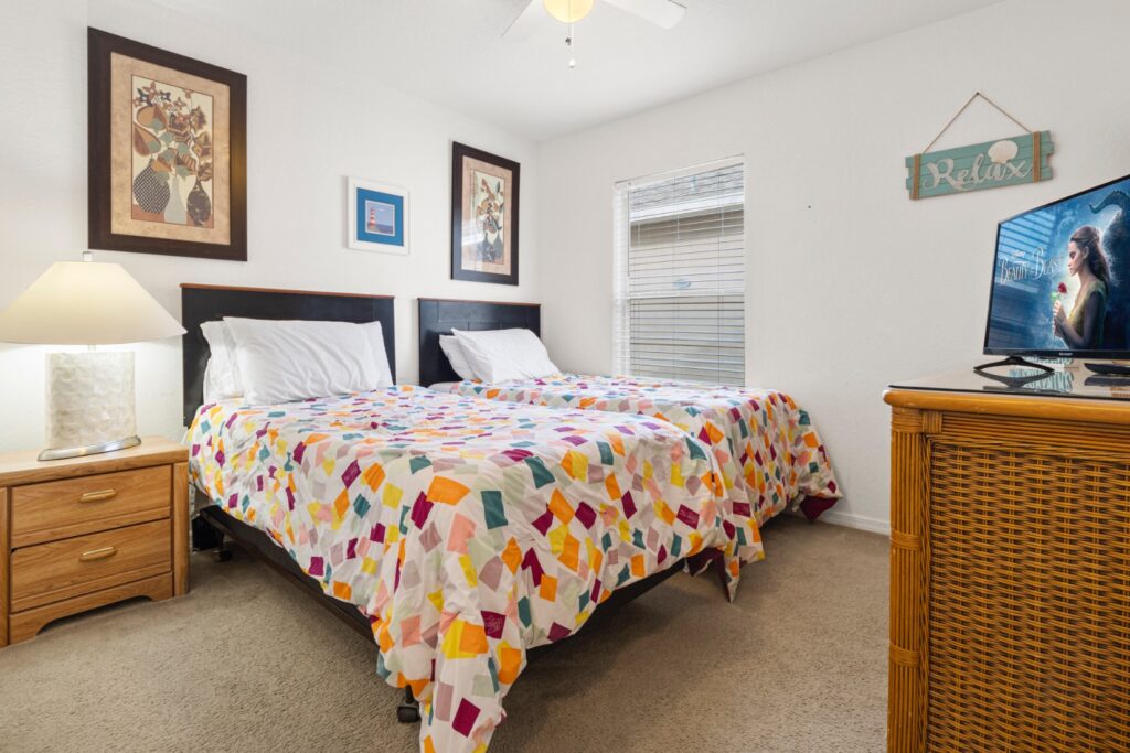 Bright twin bedroom with two beds dressed in colourful patchwork quilts, framed artwork on the walls, a bedside lamp, chest of drawers and wall-mounted TV. A 'Relax' sign adds a playful holiday touch.
