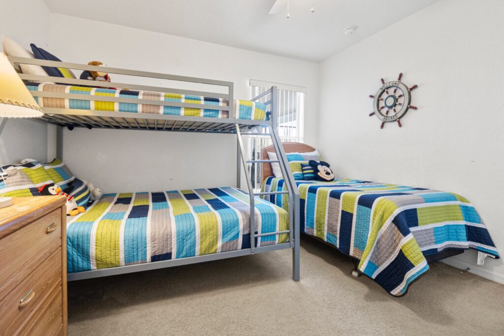 Fun children's bedroom with a metal bunk bed and a separate single bed, dressed in striped blue and green quilts with soft toys. A nautical ship's wheel wall decoration reads 'Welcome Aboard'.