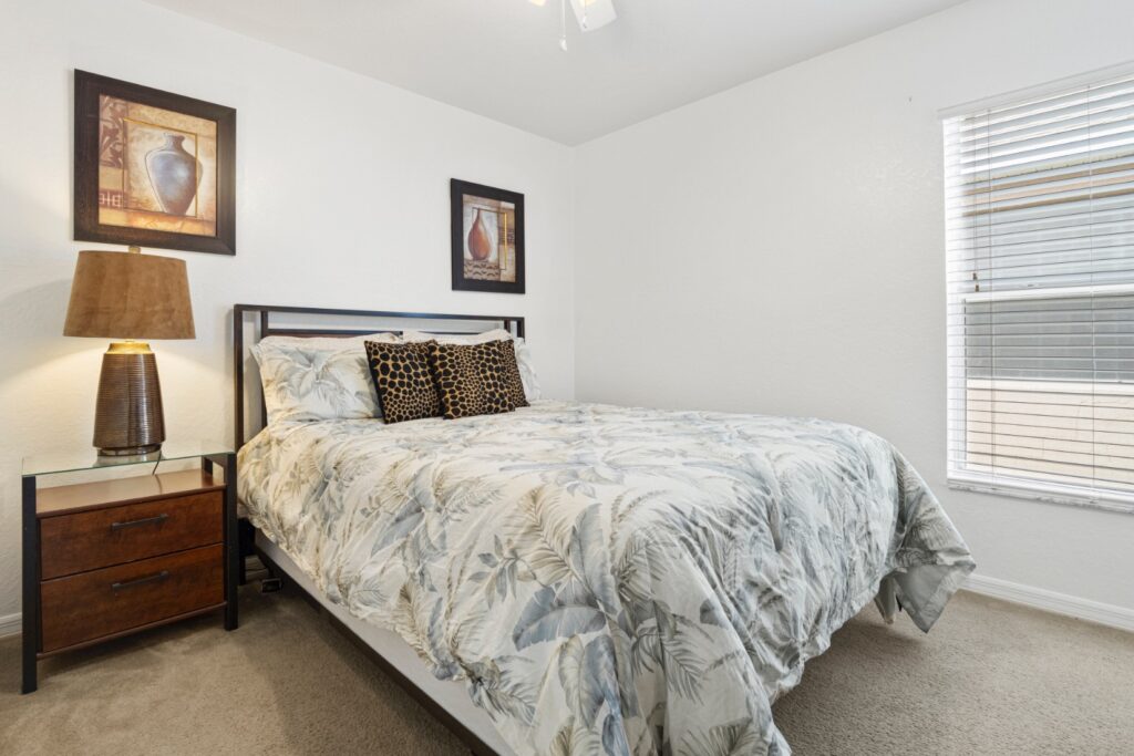 Quietly decorated queen bedroom with a metal-framed bed dressed in a botanical-print duvet, warm bedside lamp, wooden nightstand and natural light from a window with blinds.
